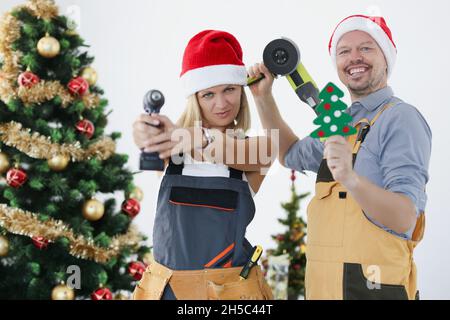 Jeune femme et homme souriant bâtisseurs en chapeaux du père noël avec des outils sur le fond de l'arbre du nouvel an. Banque D'Images