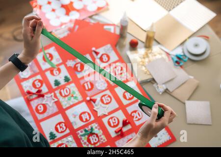 Vue de dessus d'une jeune femme méconnue utilisant un ruban de vacances pour encadrer le tableau avec des enveloppes de calendrier de l'Avent à la maison. Banque D'Images