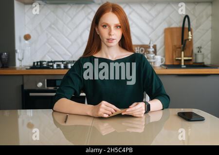 Portrait d'une jeune femme attrayante tenant dans les mains enveloppe avec lettre reçue assis à table dans la cuisine avec intérieur moderne et clair. Banque D'Images