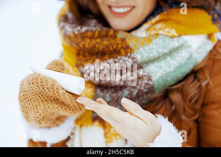 Gros plan sur la femme heureuse en chapeau marron et foulard avec des moufles utilisant la crème pour les mains dehors dans le parc de la ville en hiver. Banque D'Images