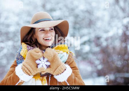 femme élégante souriante en chapeau et écharpe marron avec moufles et flocons de neige en peau de mouton en plein air dans le parc de la ville en hiver. Banque D'Images