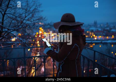 Vue de derrière une femme d'âge moyen dans un chapeau marron et une écharpe en plein air dans le parc de la ville en hiver, sous un manteau de peau de mouton en utilisant des applications de smartphone la nuit Banque D'Images