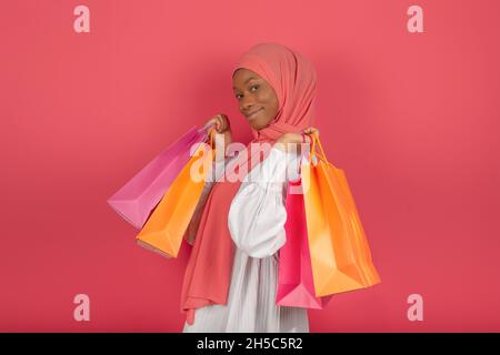 Concept de ventes saisonnières et de shopaholic.Femme musulmane souriante dans le profil tenant beaucoup de sacs lumineux de shopping regardant la caméra sur un fond rose Banque D'Images