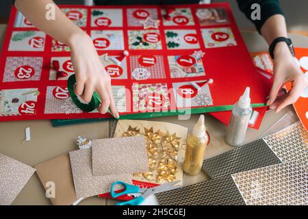 Vue en grand angle d'une jeune femme méconnue utilisant un ruban de vacances pour encadrer le tableau avec des enveloppes de calendrier de l'Avent à la maison. Banque D'Images