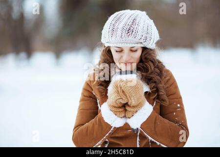 femme élégante et décontractée de 40 ans avec moufles et tasse de café dans un bonnet tricoté et un manteau de peau de mouton en plein air dans le parc de la ville en hiver. Banque D'Images