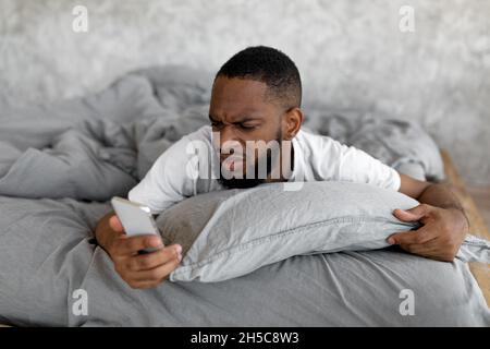 Homme noir confus regardant le téléphone dans le lit Banque D'Images
