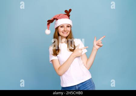 Gros plan portrait, elle est belle, charmante petite fille gaie avec des cornes de cerf en t-shirt blanc, sauvage look festif, montre un signe avec ses doigts sur le côté sur un fond bleu. Banque D'Images