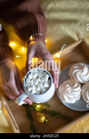 Une grande tasse de cacao dans les mains d'une fille dans un chandail, dans le fond d'une meringue et des lumières.Vue de dessus. Banque D'Images