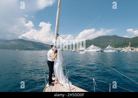 La mariée et le marié s'embrasent tout en se tenant sur l'arc d'un yacht blanc naviguant dans la baie de Kotor Banque D'Images