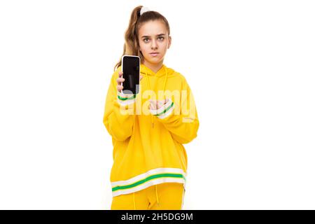 Photo d'une belle fille aux cheveux foncés avec une queue de cheval dans un élégant jaune tracksuit avec des rayures vertes montre un téléphone portable avec un blanc noir Banque D'Images