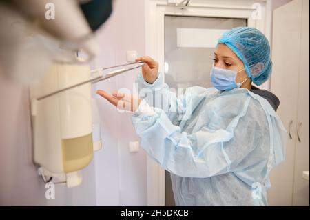 Femme chirurgien dans le masque médical et les vêtements de travail professionnels debout dans la salle de lavage de la clinique chirurgicale et utilisant le distributeur assainisseur pour la désinfection et Banque D'Images