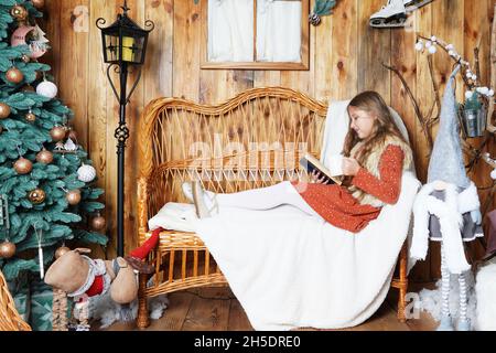 Jeune fille souriante aux cheveux longs allongé sur un canapé en osier, lisant un livre, tenant une tasse de boisson chaude près de l'arbre de Noël décoré.Fille à Noël. Banque D'Images