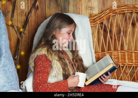 Jeune fille souriante aux cheveux longs allongé sur un canapé en osier, lisant un livre, tenant une tasse de boisson chaude près de l'arbre de Noël décoré.Fille à Noël. Banque D'Images