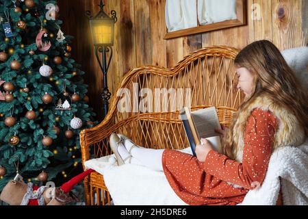 Jeune fille souriante aux cheveux longs allongé sur un canapé en osier, lisant un livre, tenant une tasse de boisson chaude près de l'arbre de Noël décoré.Fille à Noël. Banque D'Images