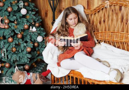 Jeune fille souriante aux cheveux longs allongé sur un canapé en osier, lisant un livre, tenant une tasse de boisson chaude près de l'arbre de Noël décoré.Fille à Noël. Banque D'Images