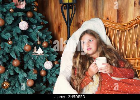 Jeune fille souriante aux cheveux longs, allongé sur un canapé en osier, rêvant, tenant une tasse de boisson chaude près de l'arbre de Noël décoré.Fille à Noël. Banque D'Images