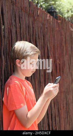 Portrait d'un garçon blond avec un t-shirt orange debout contre une clôture en bois dans la rue et à l'aide de son téléphone cellulaire Banque D'Images