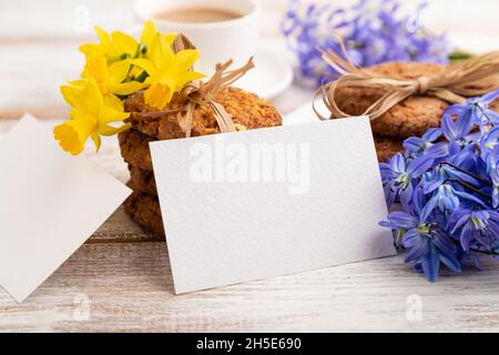 Carte de visite en papier blanc avec biscuits aux flocons d'avoine, cloches de fleurs de neige printanières, narcisses et tasse de café sur fond de bois blanc. Banque D'Images