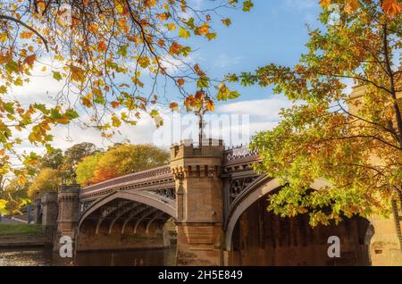 Un pont en pierre historique, orné de ferronnerie et de tours, s'étend sur une rivière.Les arbres automnaux sont au premier plan et un bleu avec des nuages ciel est au-dessus. Banque D'Images