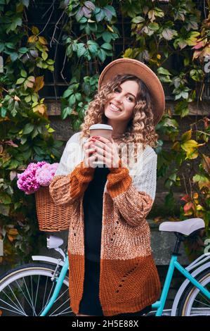 Portrait d'une jolie fille avec une expression du visage heureuse, qui boit du café à l'extérieur sur le fond du vélo de ville pour femmes qui penchait au mur trop grand par ivy. Banque D'Images