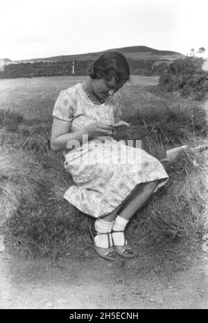 Années 1940, historique, une adolescente en robe d'été, des chaussettes de cheville et des chaussures à talons de sable, assis sur une banque herbeuse lisant un journal ou un petit livre, Angleterre, Royaume-Uni. Banque D'Images