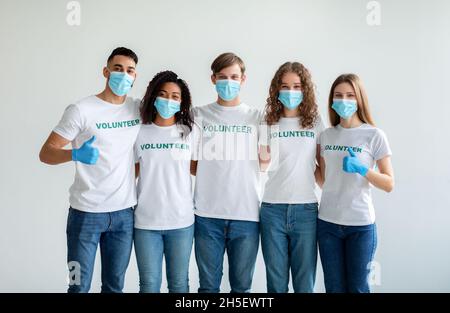 Groupe de personnes multiraciales heureuses dans des masques et des gants montrant les pouces vers le haut tout en posant sur le mur de studio léger Banque D'Images