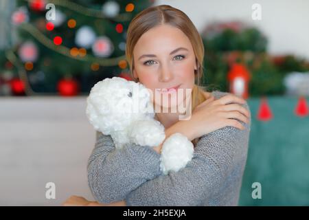 Portrait d'une belle femme qui embrasse un adorable petit ours blanc en peluche.Passer des vacances de Noël près de l'arbre de Noël à la maison.Soirée festive confortable. Banque D'Images