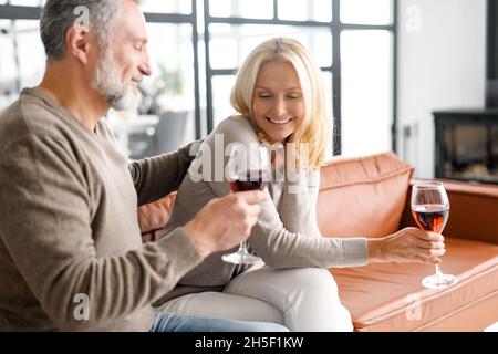 Heureux couple senior s'amuser à la maison, profiter du temps ensemble à la maison, beau homme aux cheveux gris et belle femme blonde boire du vin et rire et flirter, ont une date Banque D'Images