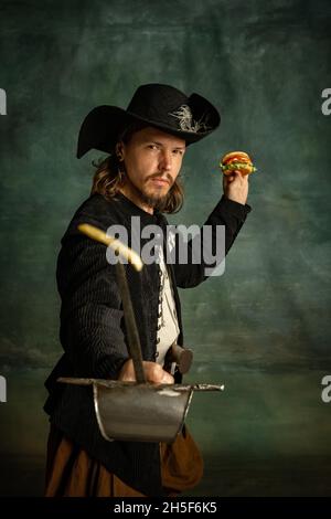 Portrait d'un homme brutal, pirate médéival tenant un hamburger isolé sur fond sombre. Banque D'Images