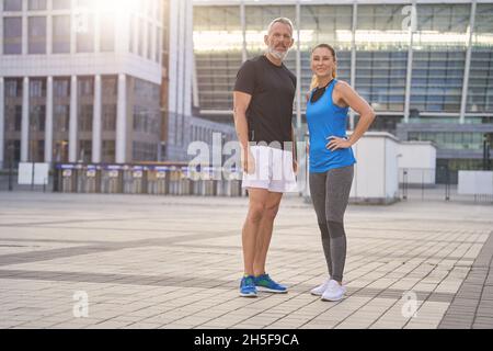 Couple actif et beau d'âge moyen dans des vêtements de sport faisant du sport ensemble dans la ville, debout à l'extérieur le matin Banque D'Images