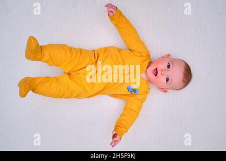 Un bébé heureux allongé sur un tapis de jeu en pyjama jaune, vue du dessus.Tout-petit souriant sur un sol blanc, espace de copie Banque D'Images