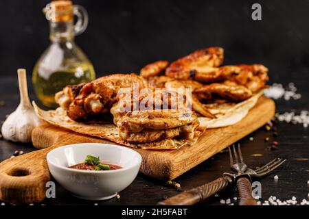 Gros plan des cuisses et ailes de poulet barbecue.Appétissant viande de volaille grillée, sauce et fourchettes sur une planche à découper en bois.Arrière-plan rustique sombre. Banque D'Images