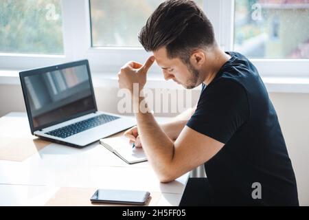 Vue latérale d'un homme d'affaires écrivant quelque chose dans son ordinateur portable à l'aide d'un smartphone mobile tout en travaillant sur un ordinateur portable avec une tablette numérique sur le bureau. Banque D'Images