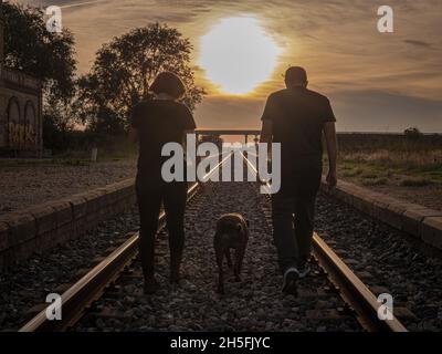 Couple adulte marchant avec leur chien sur un chemin de fer pendant le coucher du soleil Banque D'Images