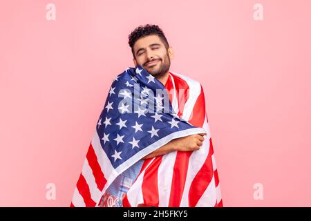 Heureux détendu satisfait beau jeune adulte barbu homme debout enveloppé dans le drapeau des États-Unis, célébrant les fêtes nationales, posant avec les yeux fermés.Studio d'intérieur isolé sur fond rose. Banque D'Images