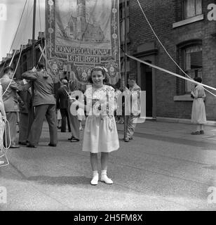 Années 1950, historique, à l'extérieur d'une rue de maisons en terrasse victorienne, une jeune fille debout pour sa photo tenant un rippon attaché à un poteau soutenant une grande couverture à la main-semée décorée pour l'église congrégationale d'Audley Range, Blackburn, Lancashire, Angleterre, Royaume-Uni.Depuis les années 1840, la révolution industrielle et la croissance des industries du coton et du textile ont transformé la zone et le logement en terrasses, les écoles, les magasins, les pubs et les églises ont été construits sur des terres agricoles pour servir la nouvelle population travaillant dans les usines et les usines de textile. Banque D'Images