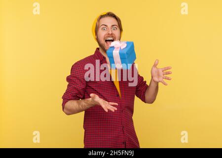 Portrait d'un gars à barbe taille basse en bonnet beanie et en chemise à carreaux, debout avec la bouche ouverte et les mains levées, essayant d'attraper des cadeaux de noël volants.Studio d'intérieur isolé sur fond jaune. Banque D'Images