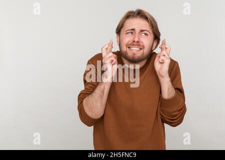 Portrait de l'homme plein d'espoir drôle avec barbe portant un sweat-shirt, croisant les doigts pour la chance, faisant un souhait, rêvant de l'intérieur, rituel.Prise de vue en studio isolée sur fond gris. Banque D'Images