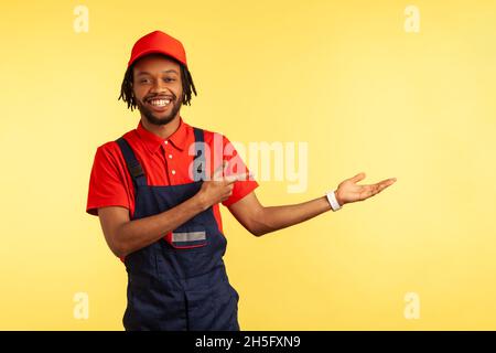 Un mécanicien barbu souriant portant une combinaison présentant un espace de copie sur la paume, pointant vers l'extérieur, faisant la publicité de la livraison ou du service d'entretien ménager.Studio d'intérieur isolé sur fond jaune. Banque D'Images