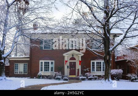 Belle maison en brique avec baies vitrées avec arbre de Noël montrant à travers et décoré des piliers et traîneau sur le porche dans la neige encadrée par des arbres d'hiver Banque D'Images