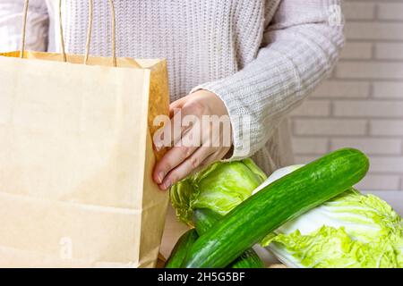 Les mains de femme caucasienne portent un sac de papier d'artisanat avec des légumes verts de la livraison en ligne de shopping à la maison dans la cuisine sur fond clair. Banque D'Images