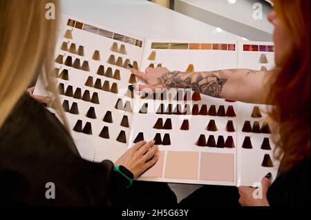 Vue arrière d'une femme coiffeuse tête rouge tatouée main, sur un catalogue de la palette de couleurs de coloration de cheveux et conseiller client, salon de coiffure client à choo Banque D'Images