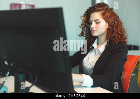 Jeune femme réussie à cheveux rouges dans un costume d'affaires et des lunettes travaillant dans un bureau sur un ordinateur Banque D'Images