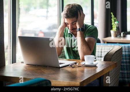 Portrait de l'homme fatigué épuisé indépendant portant un T-shirt vert, souffrant de migraine ou d'hypertension tout en travaillant en ligne sur un ordinateur portable, prise de vue en intérieur près d'une grande fenêtre, arrière-plan de café. Banque D'Images