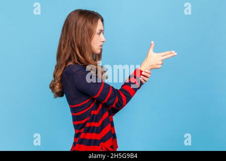 Vue latérale d'une femme en colère imitant des armes, en filant avec des pistolets à doigts, avec un aspect inquiétant., portant un pull rayé de style décontracté.Studio d'intérieur isolé sur fond bleu. Banque D'Images