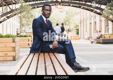 Portrait d'un homme d'affaires afro-américain noir en costume, lunettes et écouteurs assis sur un banc dans une place de ville à l'extérieur Banque D'Images