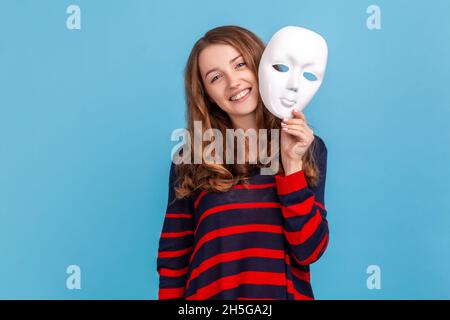 Femme souriante portant un chandail rayé de style décontracté, tenant le masque blanc, montrant son expression positive, prétendant être une autre personne.Studio d'intérieur isolé sur fond bleu. Banque D'Images