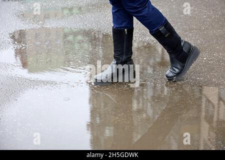 Pluie en ville, jambes de femme en bottes dans une rue avec des flaques.Les bâtiments résidentiels se reflètent dans l'eau, la femme marchant dans le temps d'automne Banque D'Images