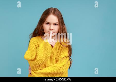 Portrait d'une petite fille sérieuse faisant le geste de silence avec le doigt sur les lèvres, regardant l'appareil photo, portant jaune style décontracté pull.Studio d'intérieur isolé sur fond bleu. Banque D'Images