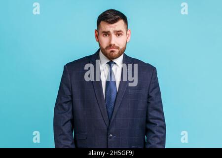 Beau homme d'affaires malheureux portant un costume de style officiel sombre regardant l'appareil photo avec une triste expression du visage, ayant des problèmes.Studio d'intérieur isolé sur fond bleu. Banque D'Images